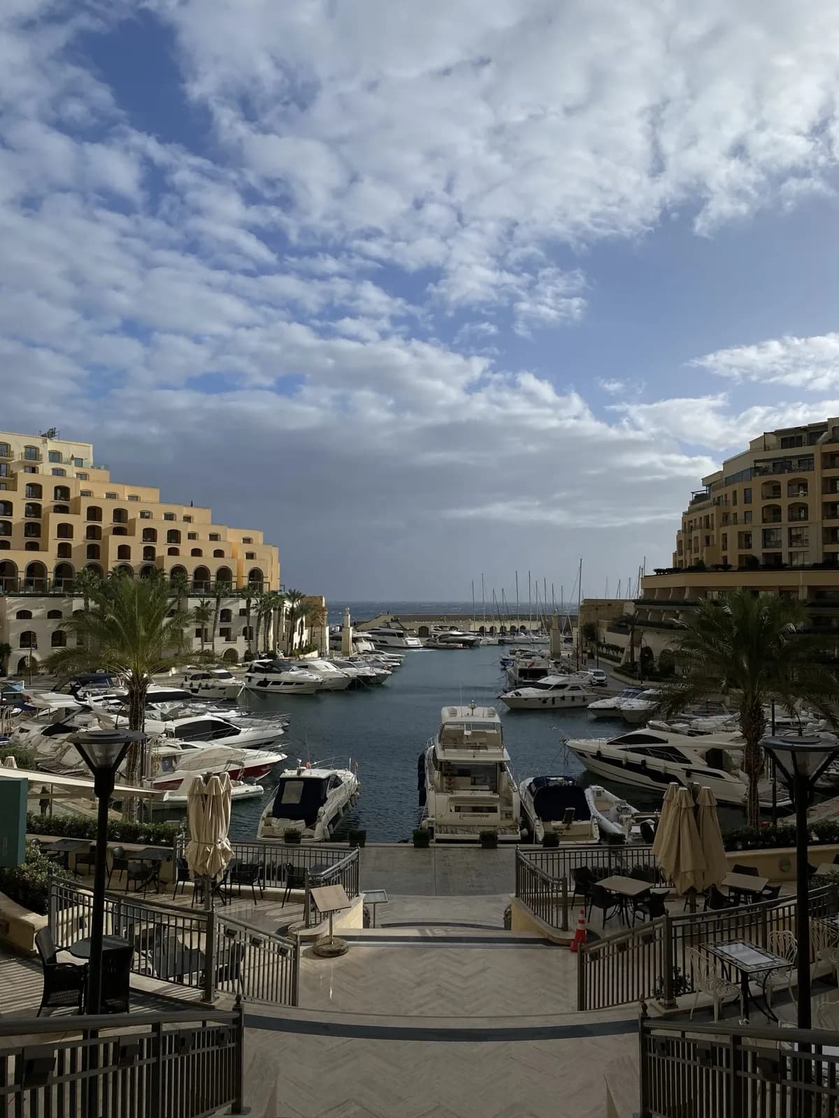 Yachts moored in Portomaso marina, framed by apartment blocks and palm trees under a cloudy sky.