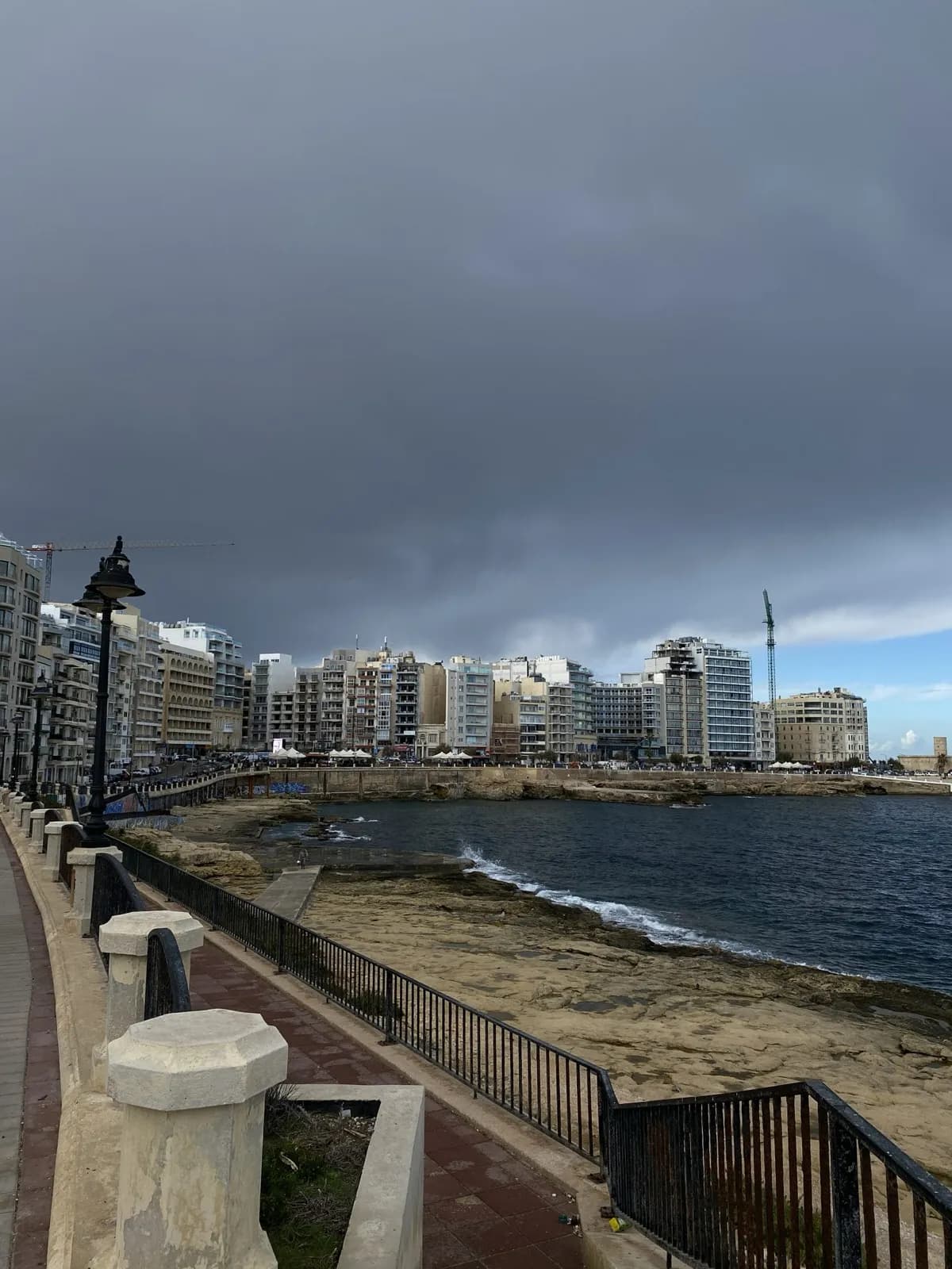 The Sliema seafront promenade with apartment blocks and a construction crane under a dark storm sky.