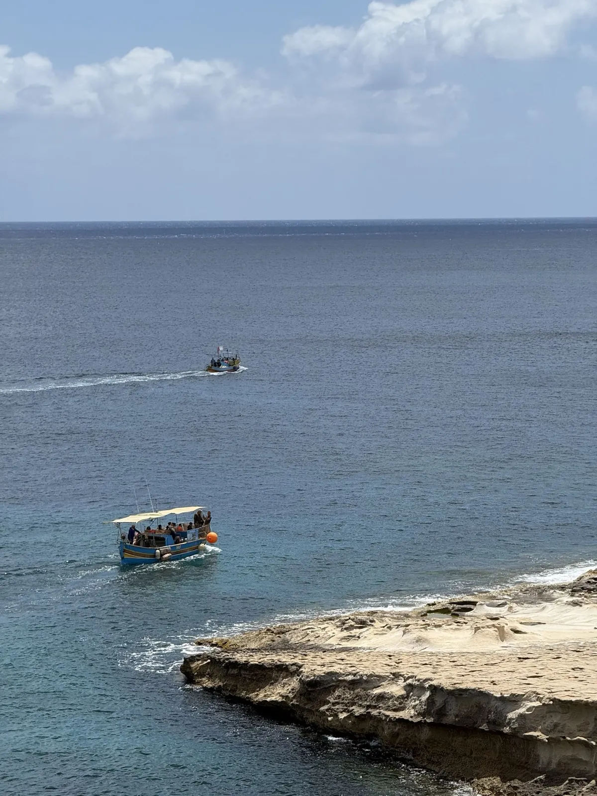 Two small boats on open blue sea off a flat limestone shelf on the Maltese coast, the nearer one carrying passengers under a canopy.
