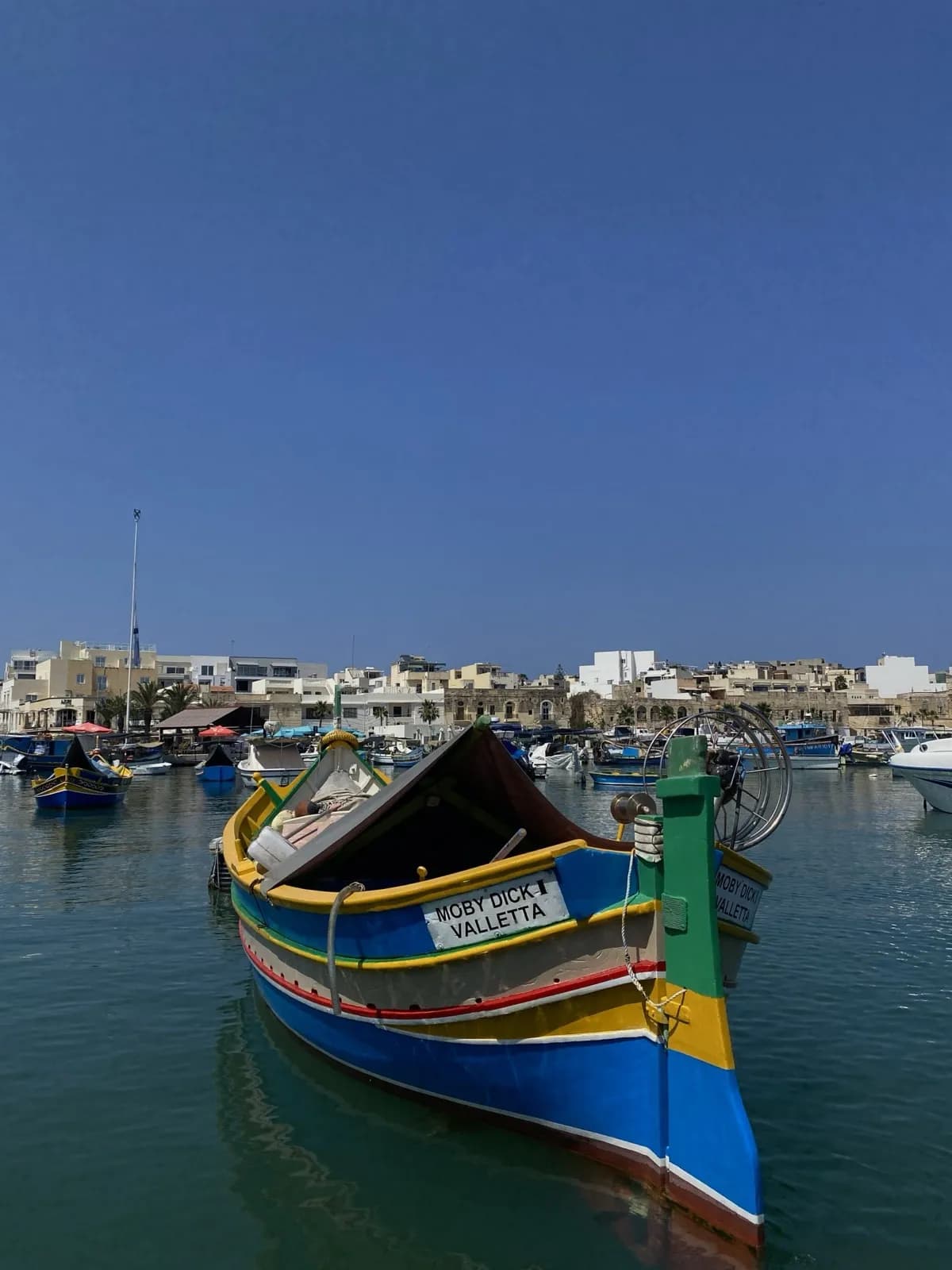A brightly painted luzzu fishing boat moored in Marsaxlokk harbour, with the village and other boats behind it